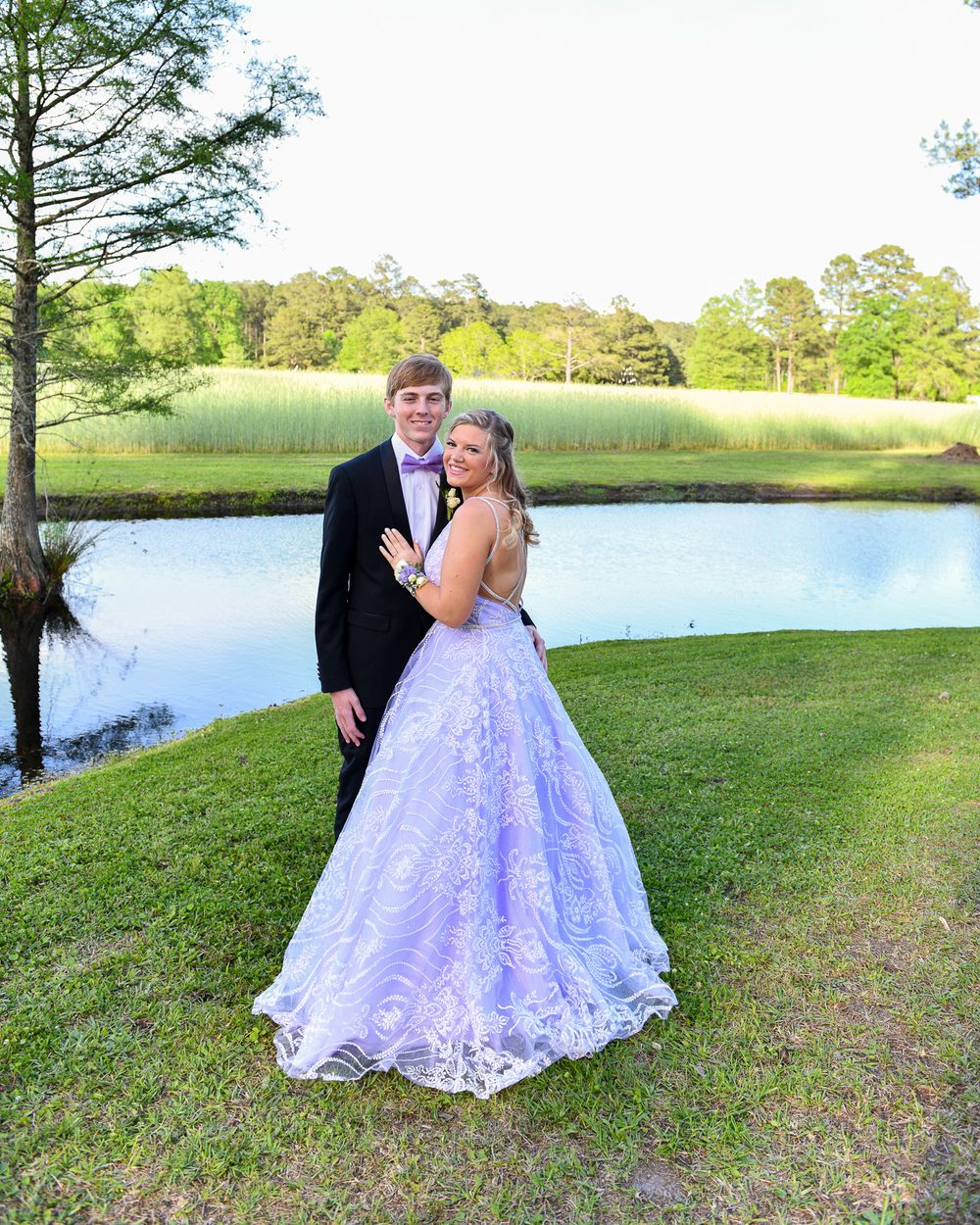 A couple poses in formal attire by a lake, with her in a lavender ball gown and him in black tuxedo.