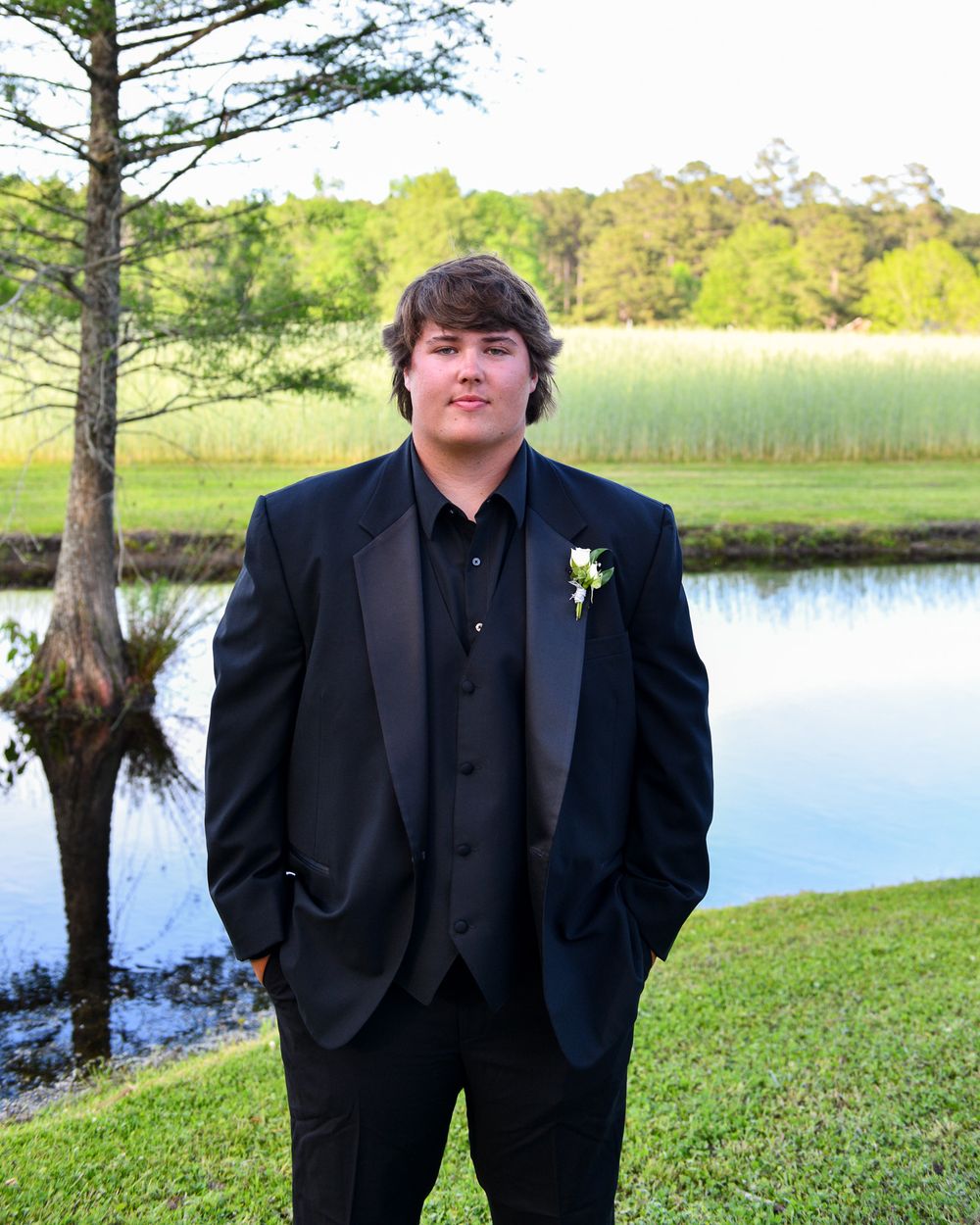 A person in a black tuxedo poses for a formal photo by a scenic lake and pine tree.