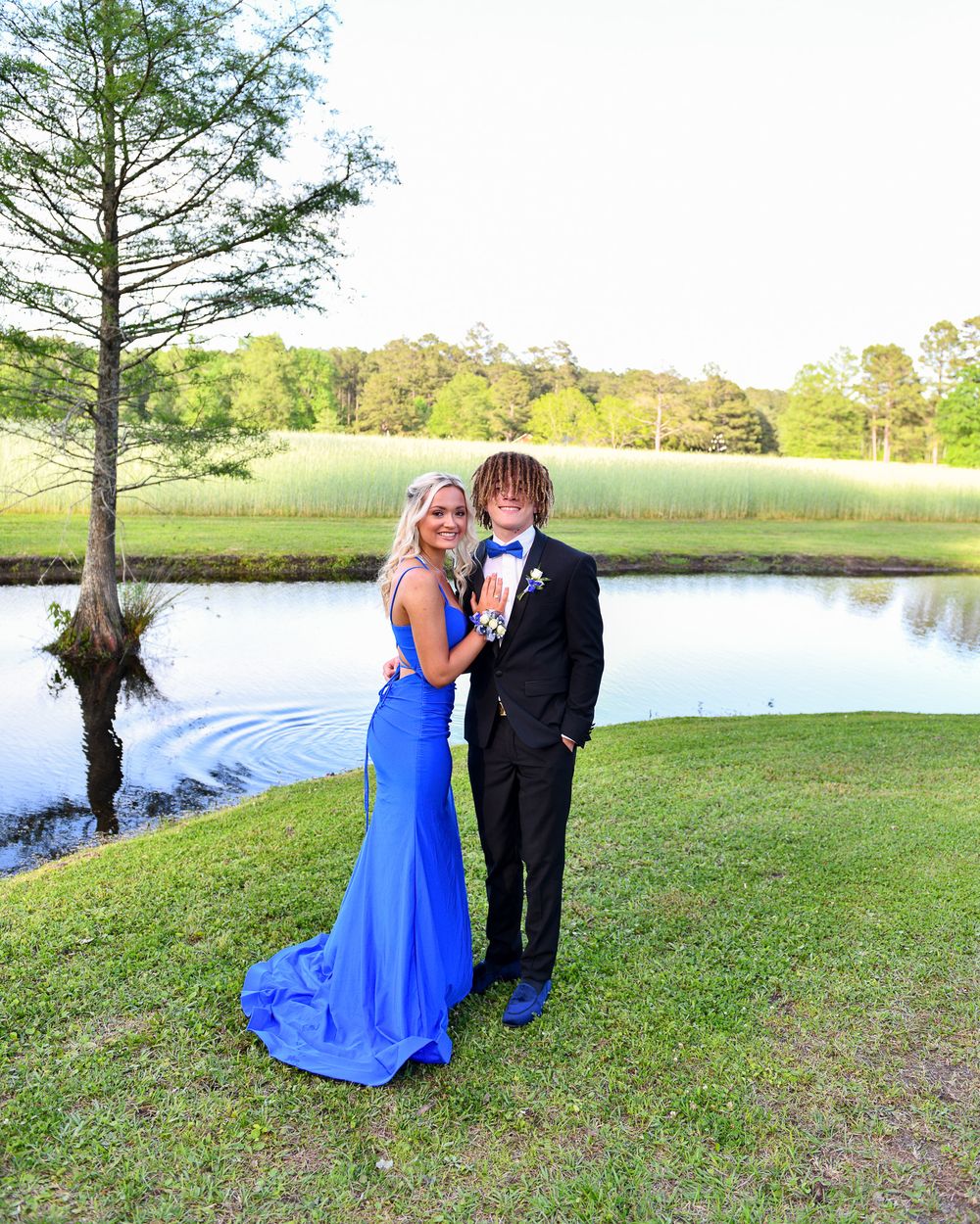 A couple in formal wear poses by a lake, with her in a royal blue gown and him in black suit.