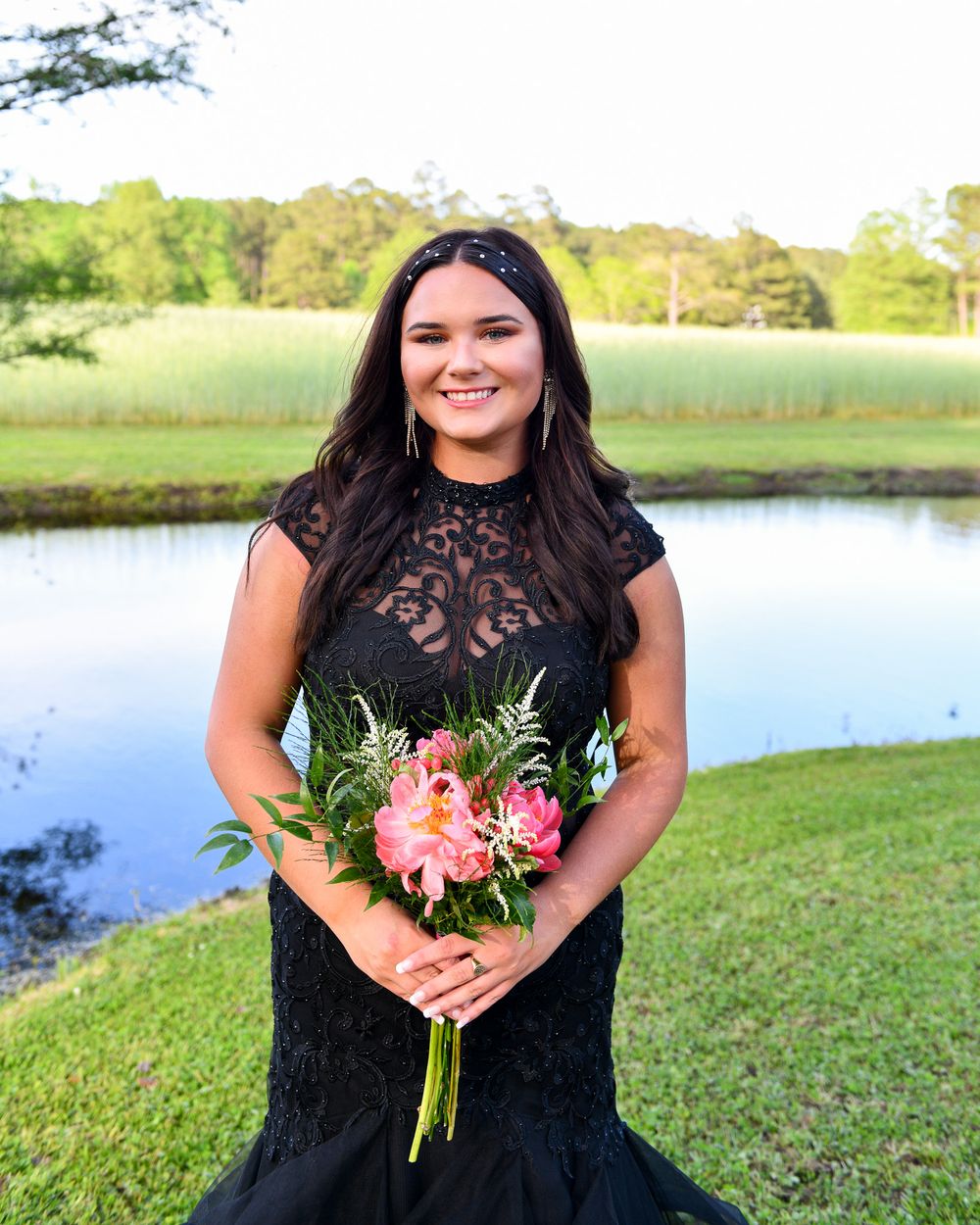 A person in a black lace dress holds coral flowers while posing by a scenic lake.