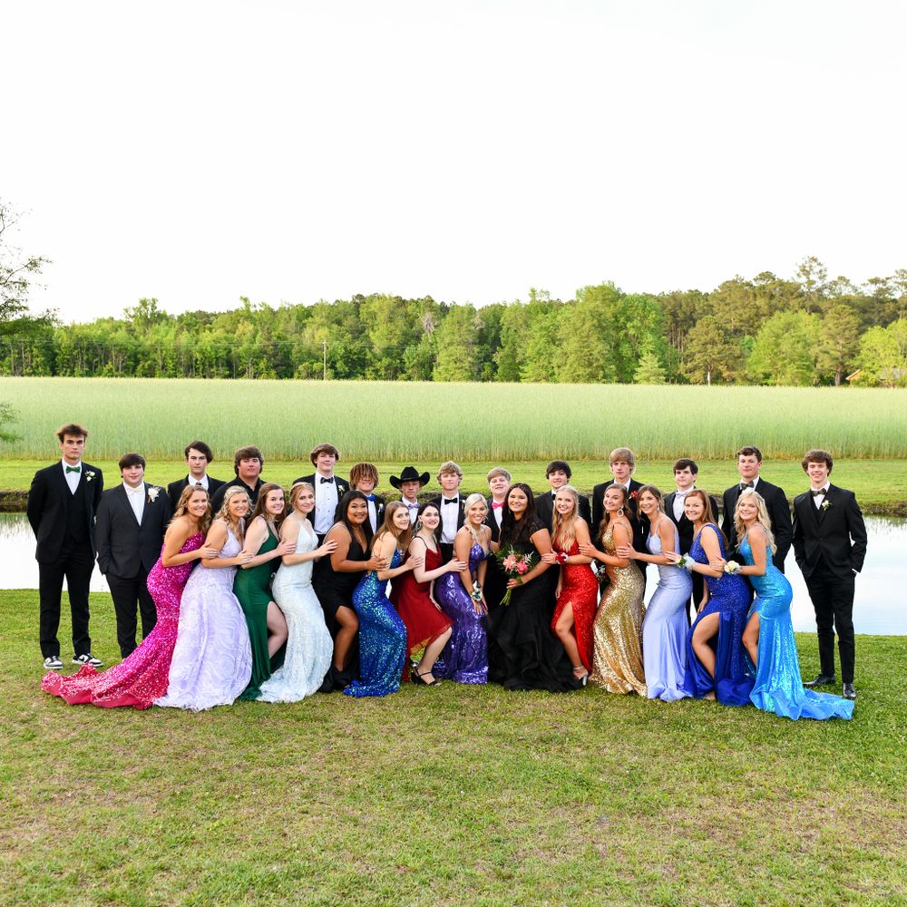 Large prom group poses together outdoors wearing colorful formal dresses and suits near a scenic field.