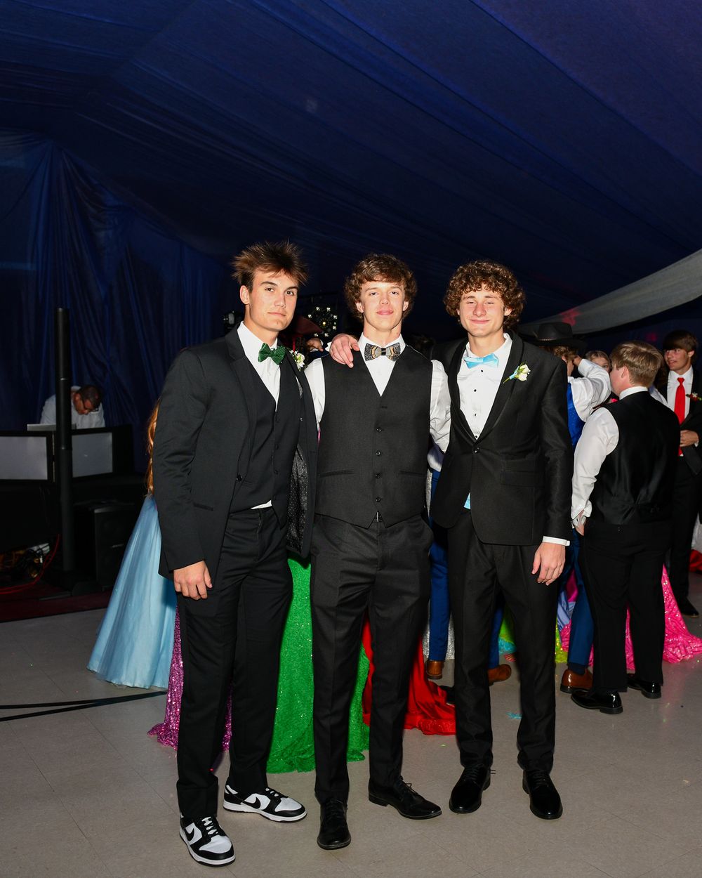 Three friends in formal suits pose together at an evening event under blue tent lighting.
