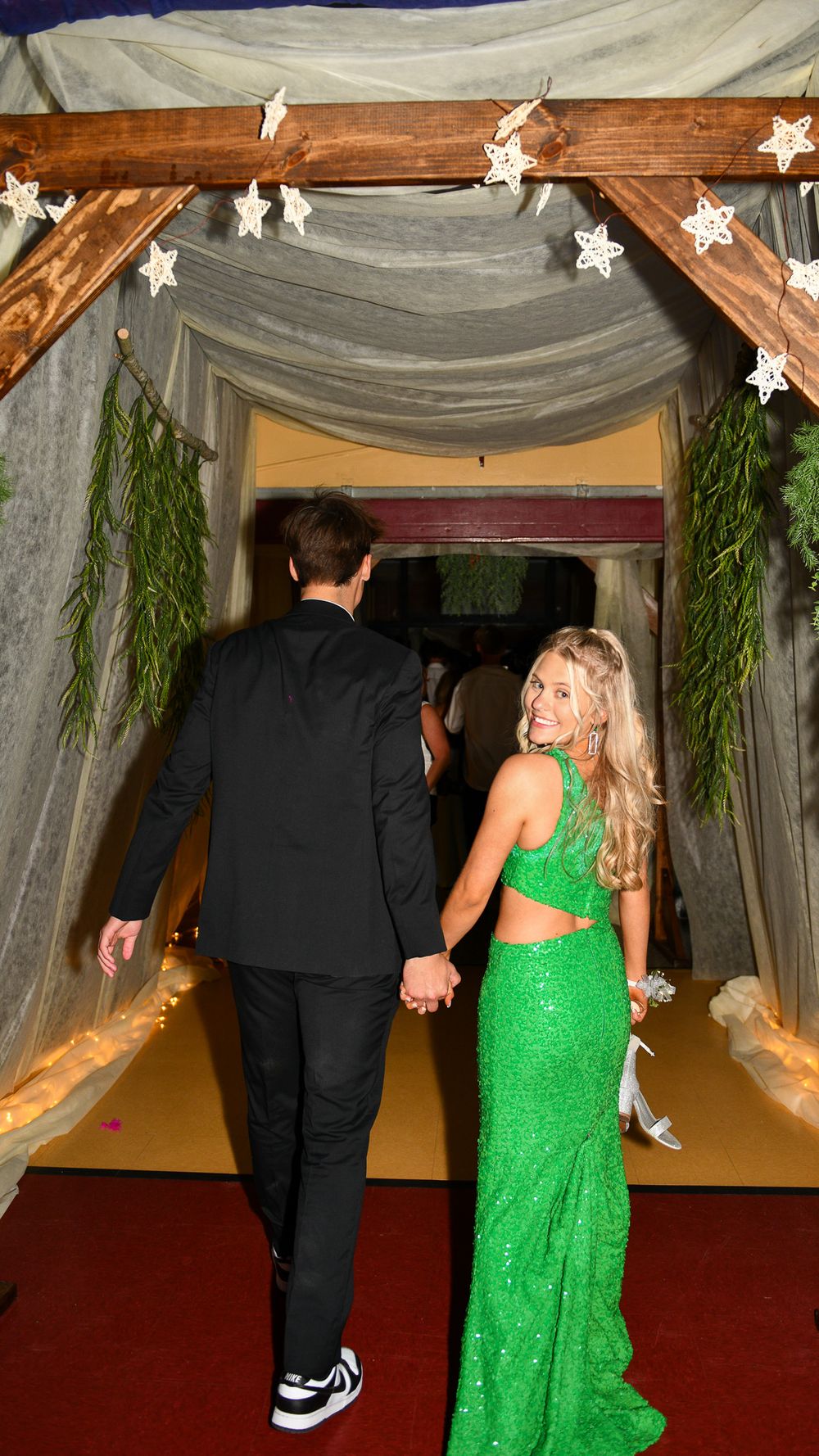 Couple walks through decorated archway with fairy lights and greenery at formal event.