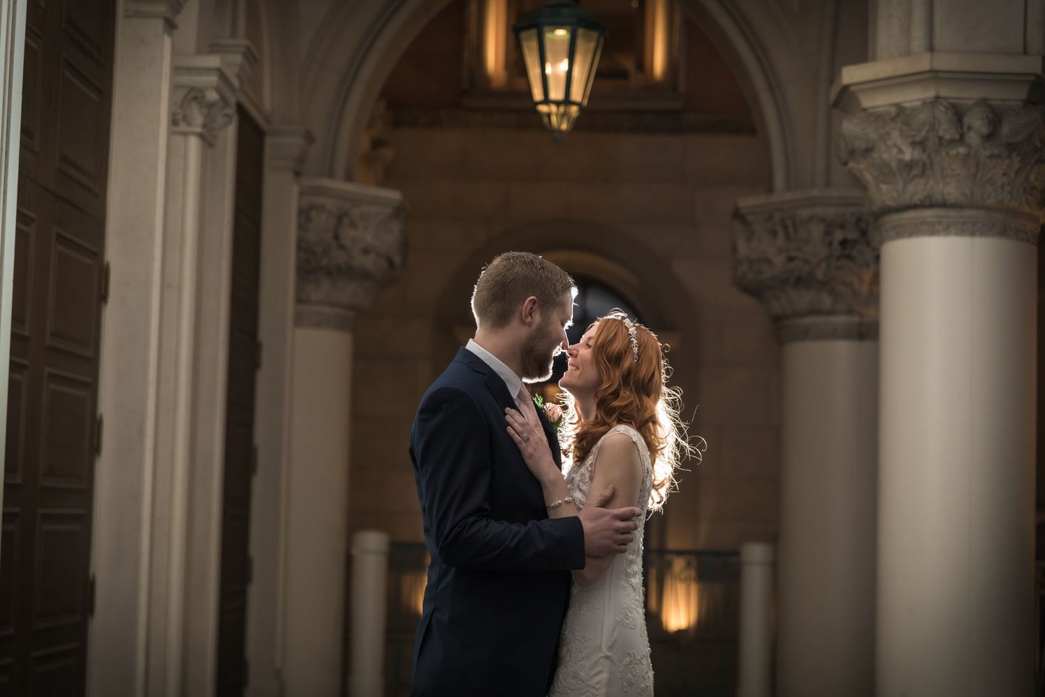 Couple shares romantic moment under elegant archway with ornate columns and warm lighting in classic wedding photo.