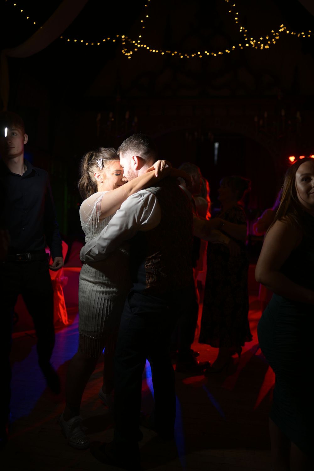 Silhouetted couples dance together in a dimly lit reception hall with string lights overhead.