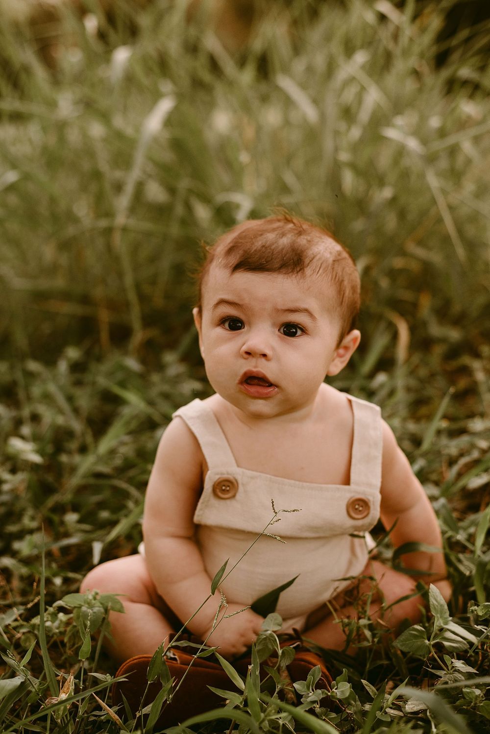Baby in tan overalls sits in grass during outdoor photo session.