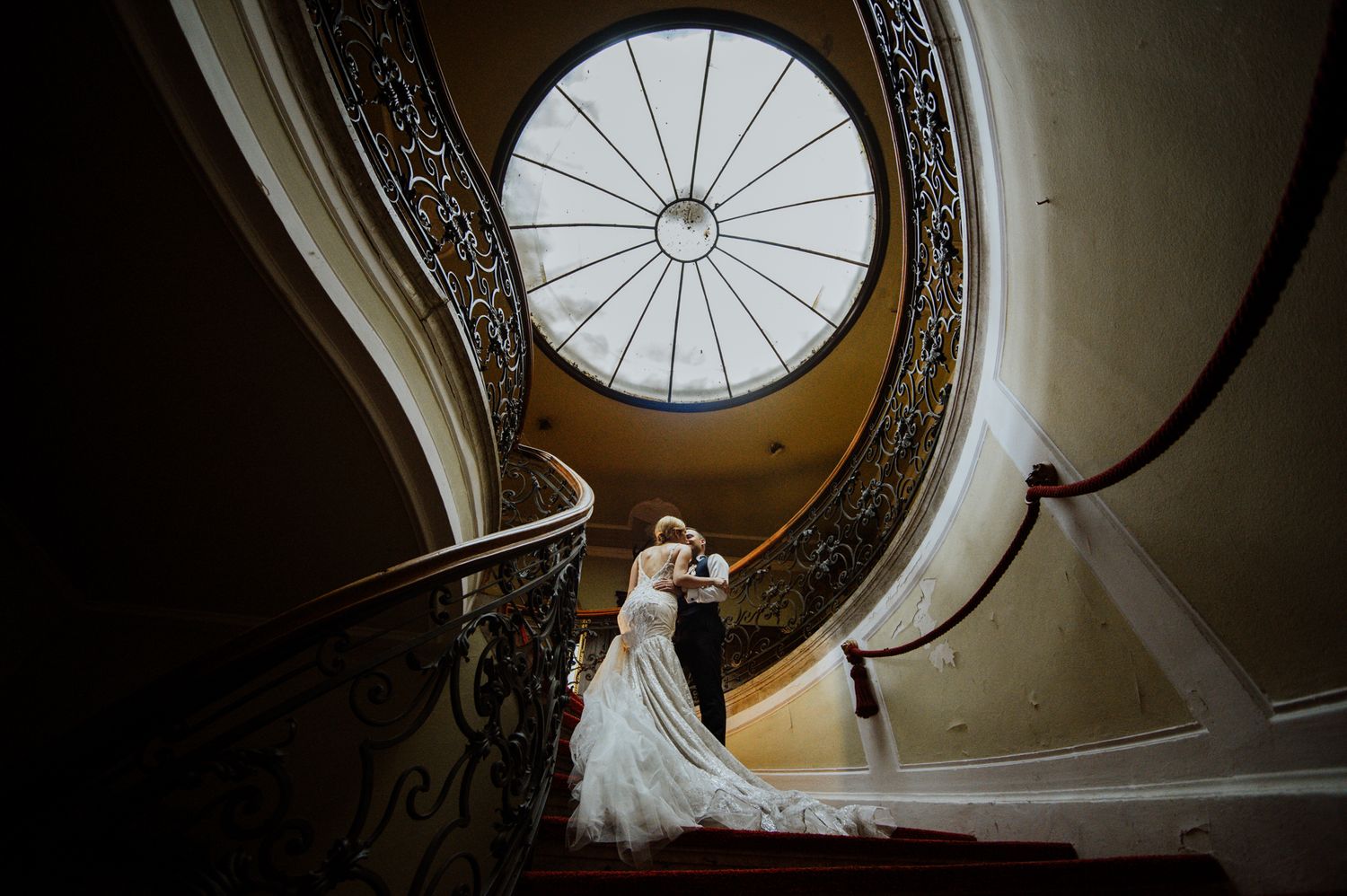 Elegant wedding photograph on spiral staircase with dramatic circular skylight and ornate railings.