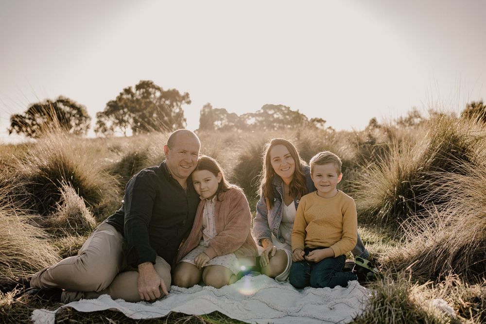 Group sitting together on blanket in grassy field during golden hour sunset with trees in background.