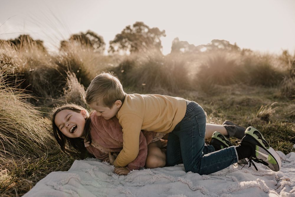Playful candid moment between children laughing and hugging on a white blanket outdoors.
