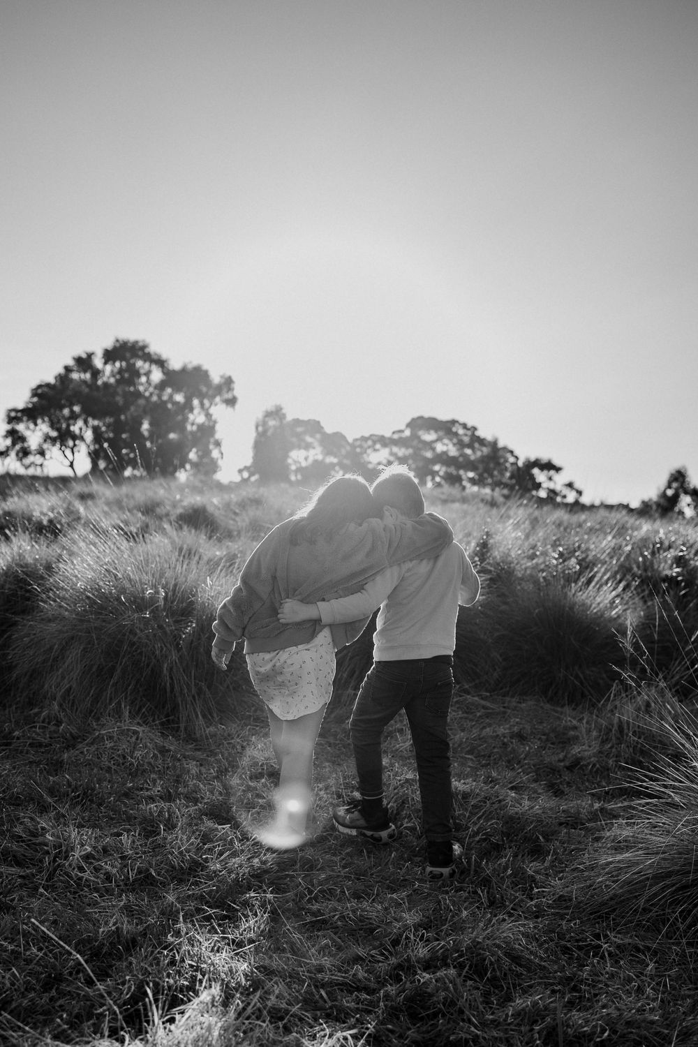 Black and white silhouette of a couple walking together through tall grass at sunset.