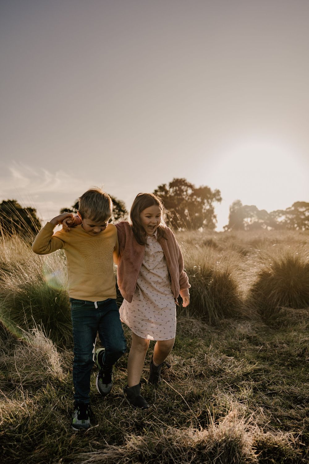 Two figures playfully walking through tall grass during golden hour, backlit by warm sunlight.