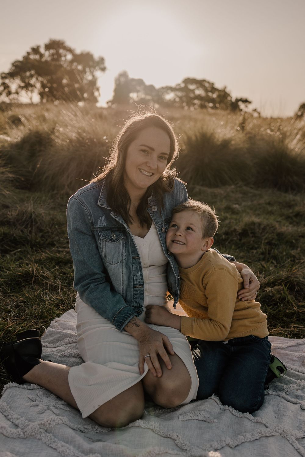 A tender sunset moment as two people share a laugh while sitting on a picnic blanket in a grassy field.
