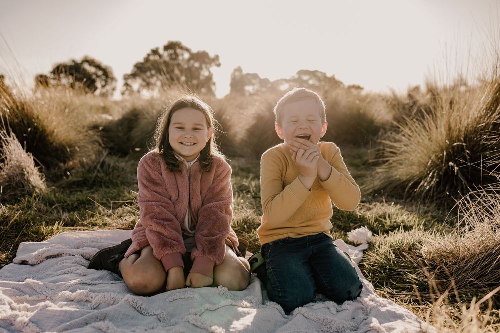 Two siblings sitting together on a blanket outdoors during golden hour with ornamental grass in the background.