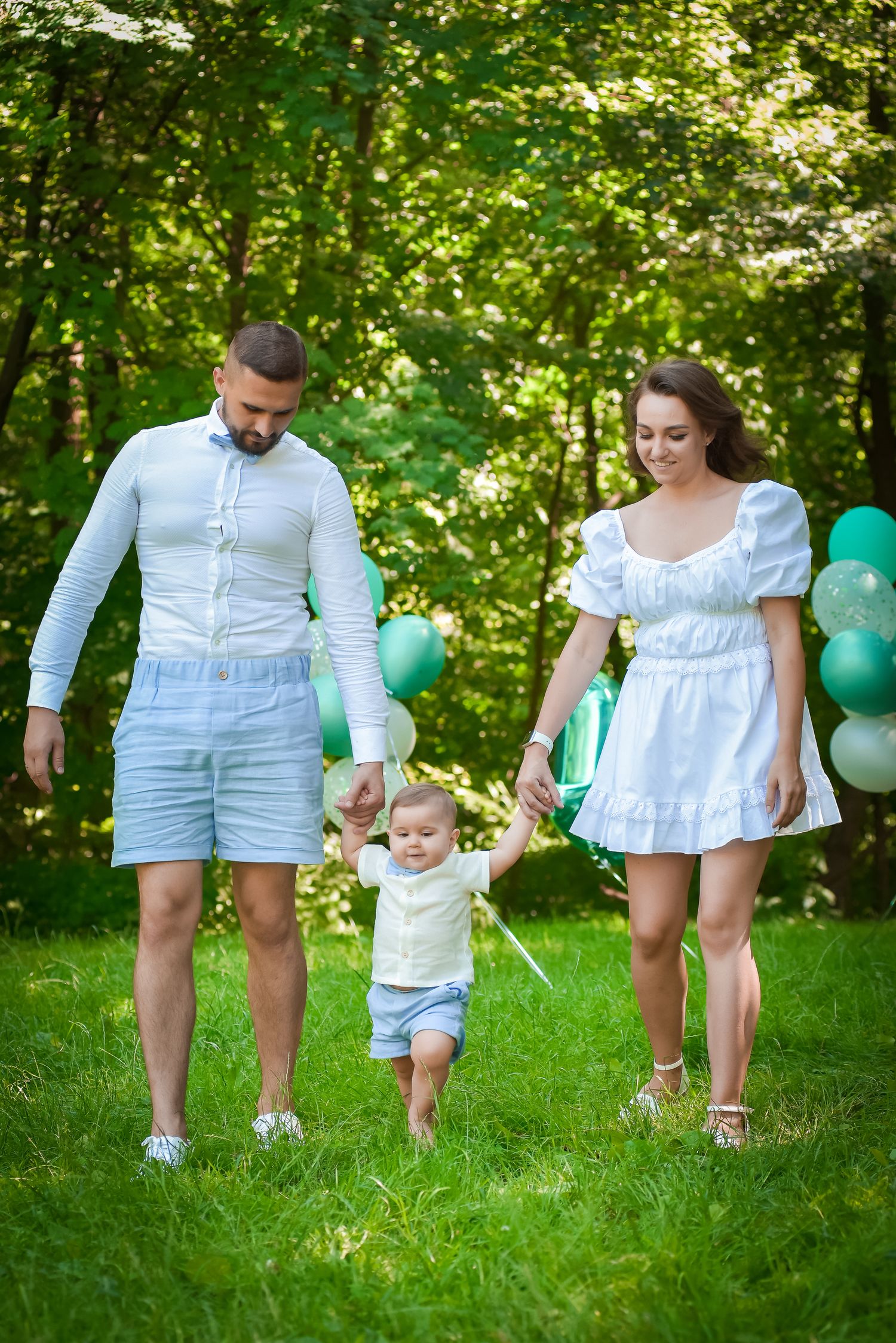Family walking together through green grass with teal balloons in summer park.