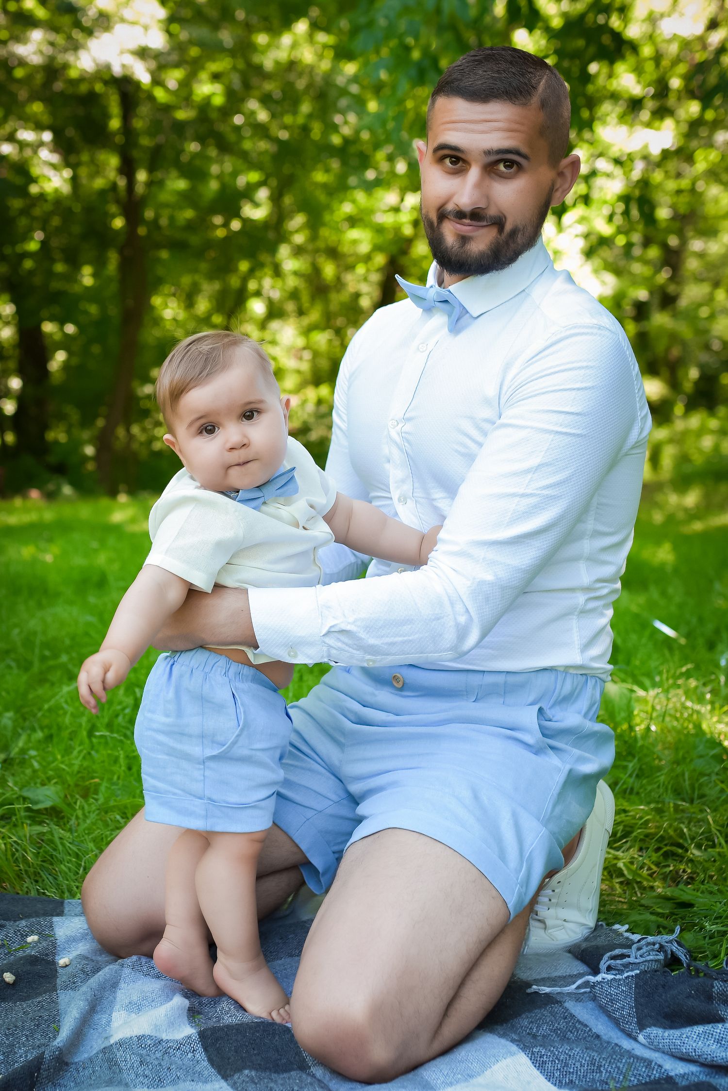 Parent and baby in white outfits sit together on a blanket in a sunny garden setting.