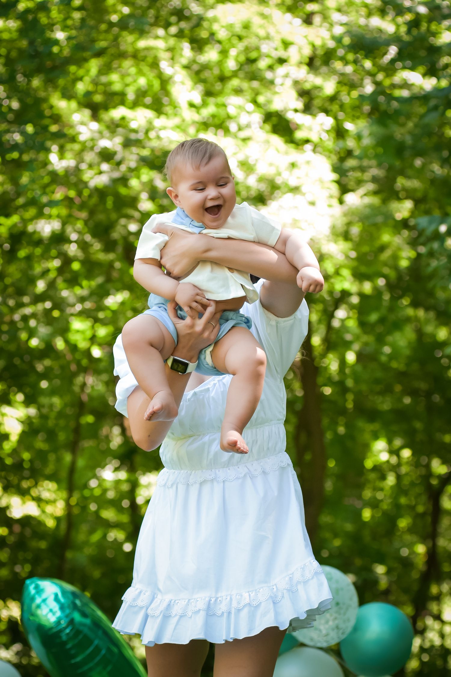 Baby being lifted playfully in the air against a green outdoor background.