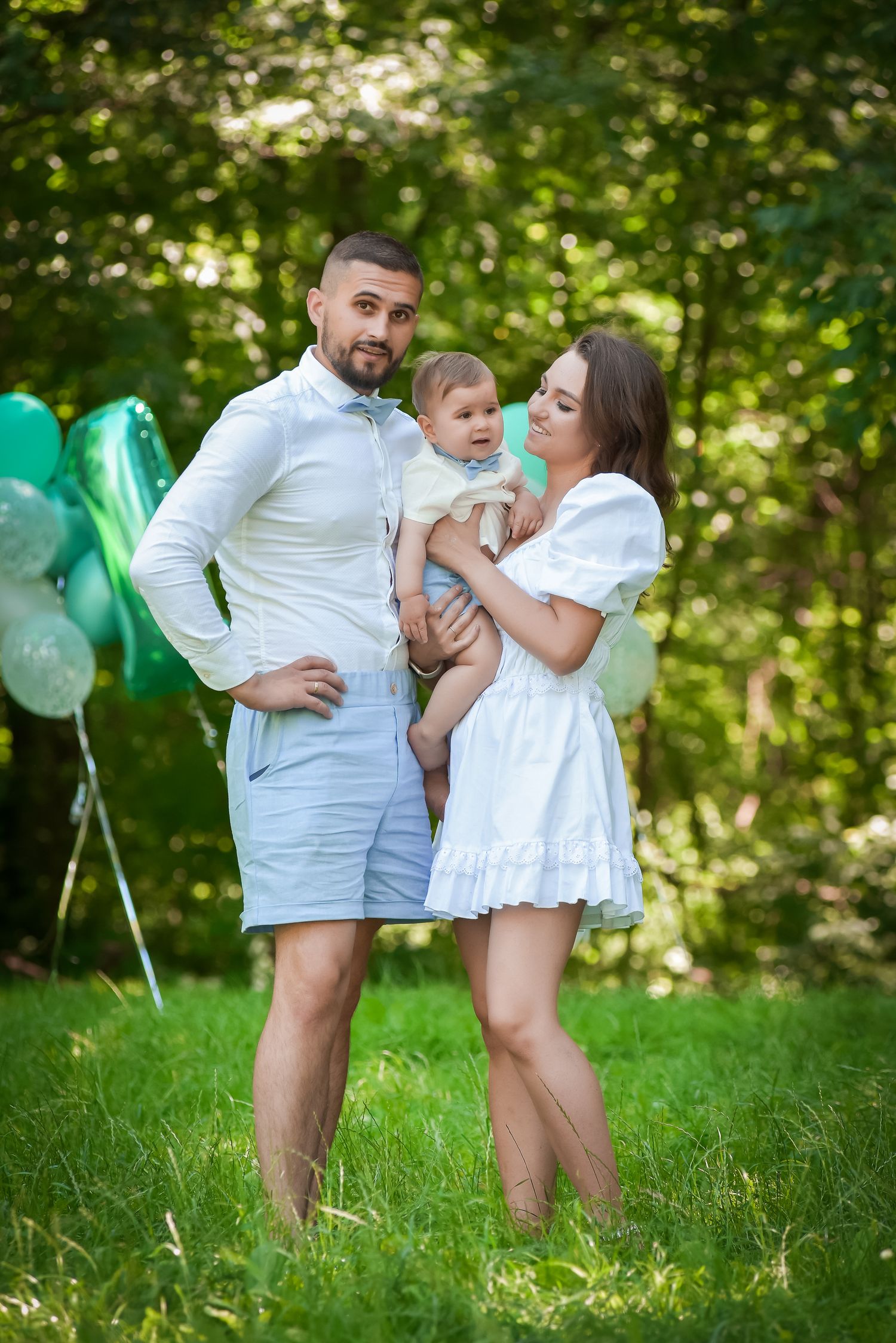 Family dressed in white playing together on grass with teal balloons in background.