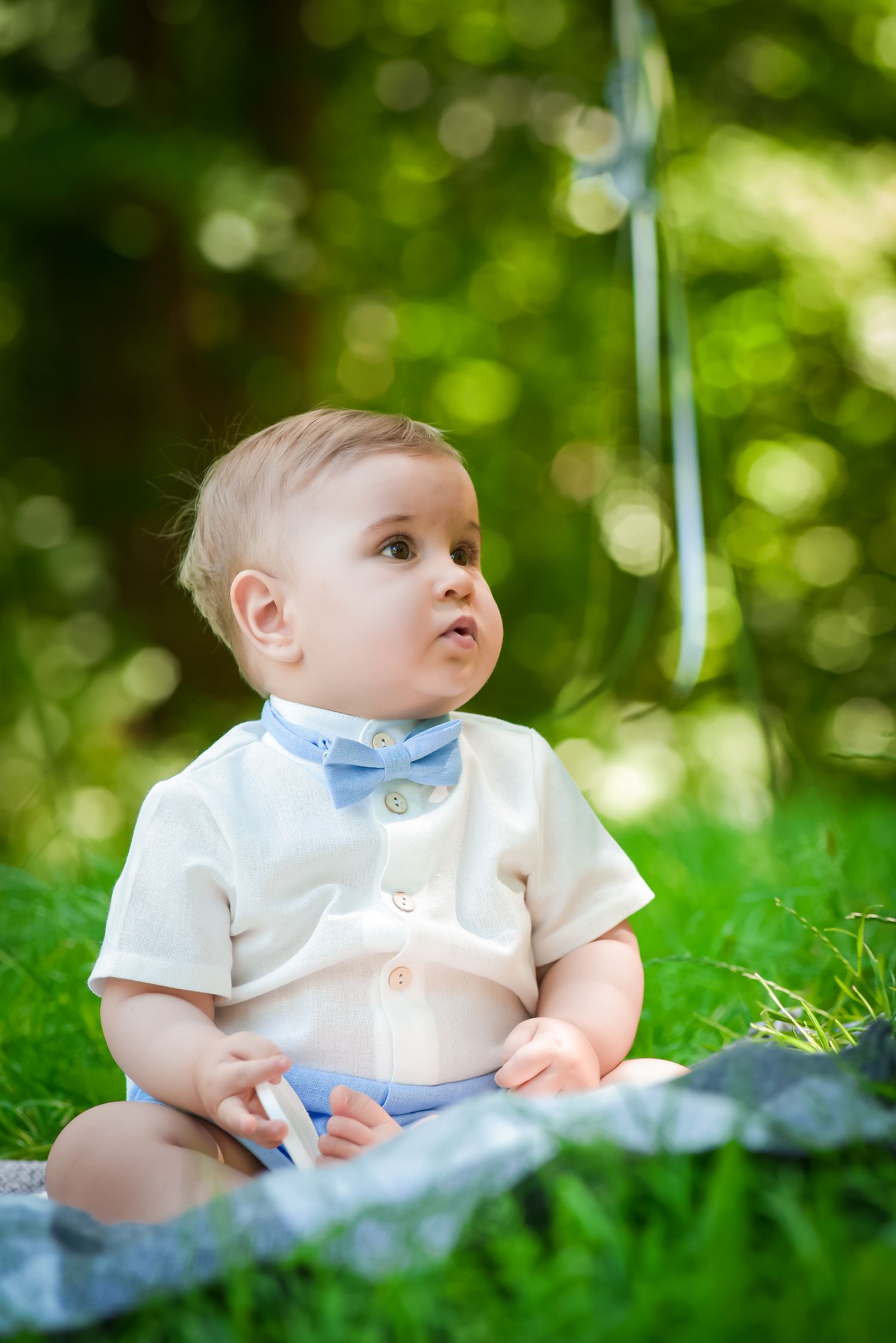 Baby in formal white and blue outfit sits on blanket in garden setting.