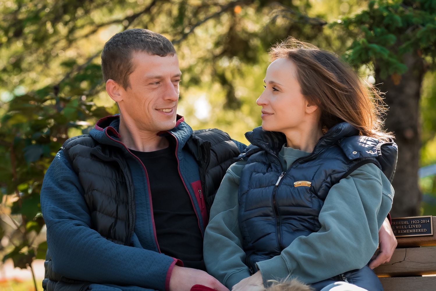 A couple in winter jackets share a tender moment while sitting outdoors in natural lighting.