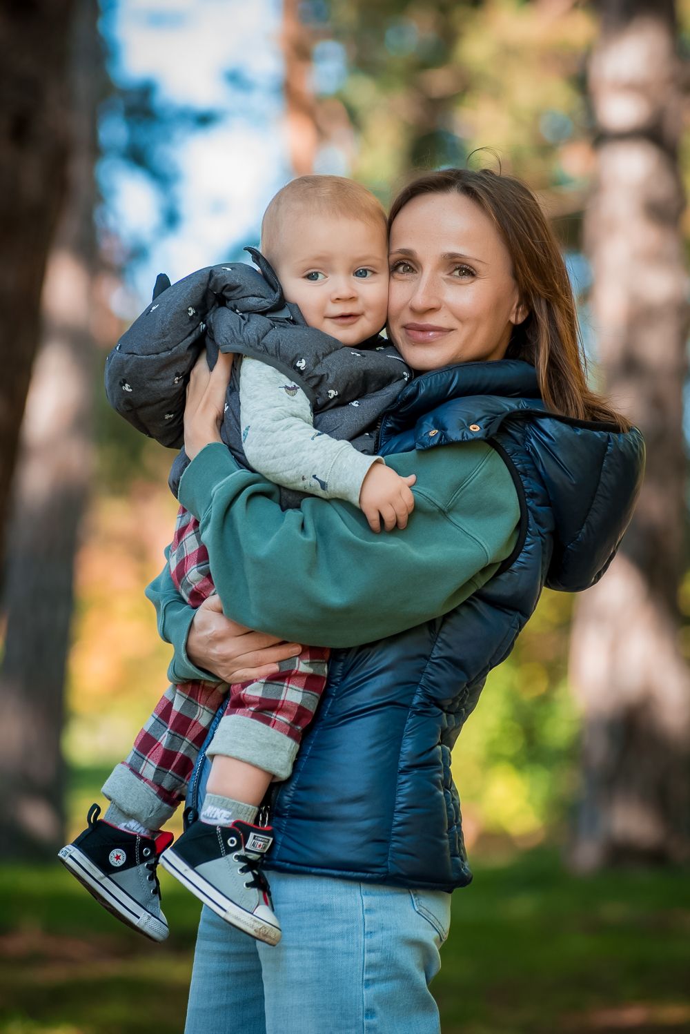 A parent in a teal jacket holds a smiling toddler wearing plaid pants and sneakers in a wooded setting.