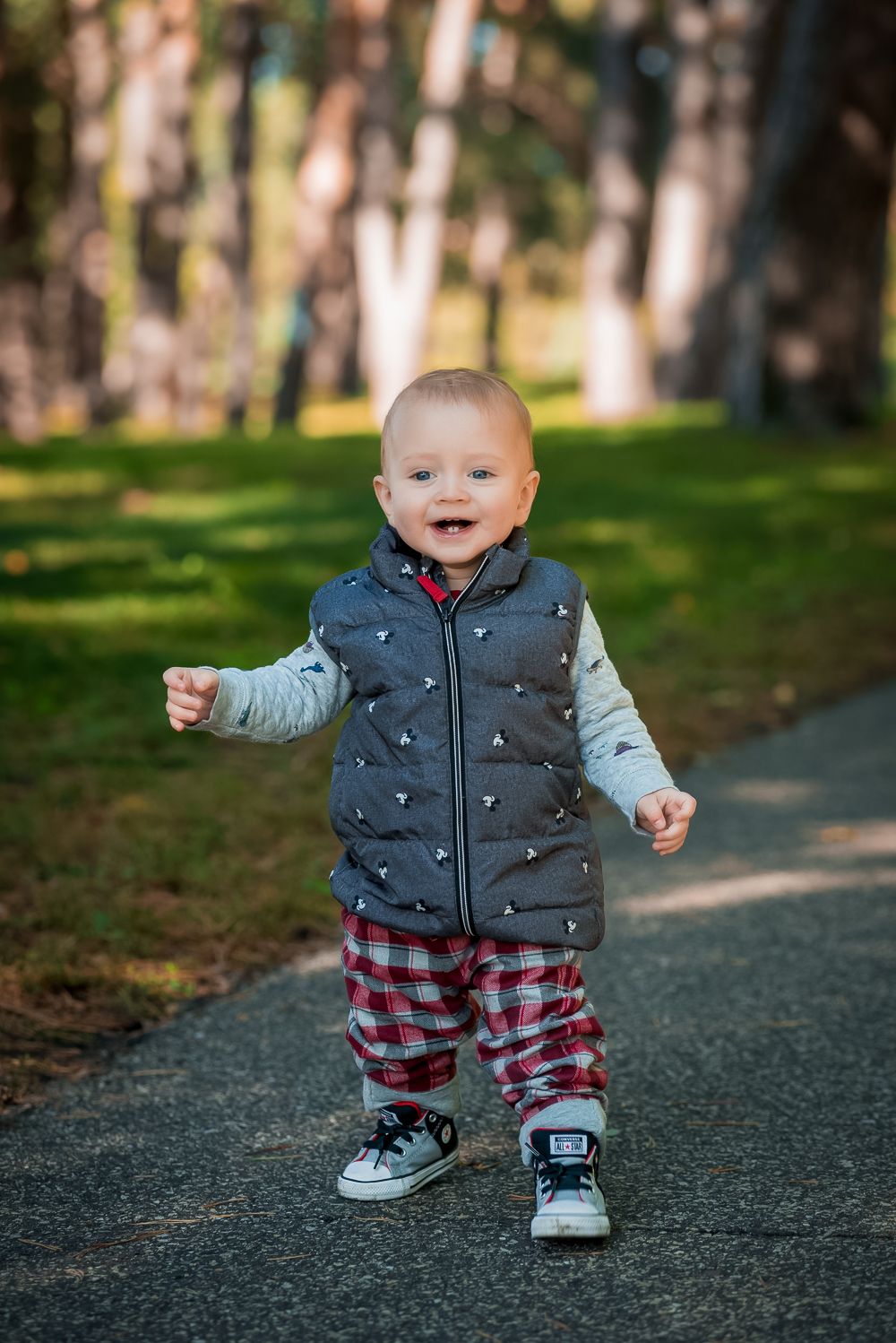 A happy toddler in a puffy vest and plaid pants takes first steps on a forest path.