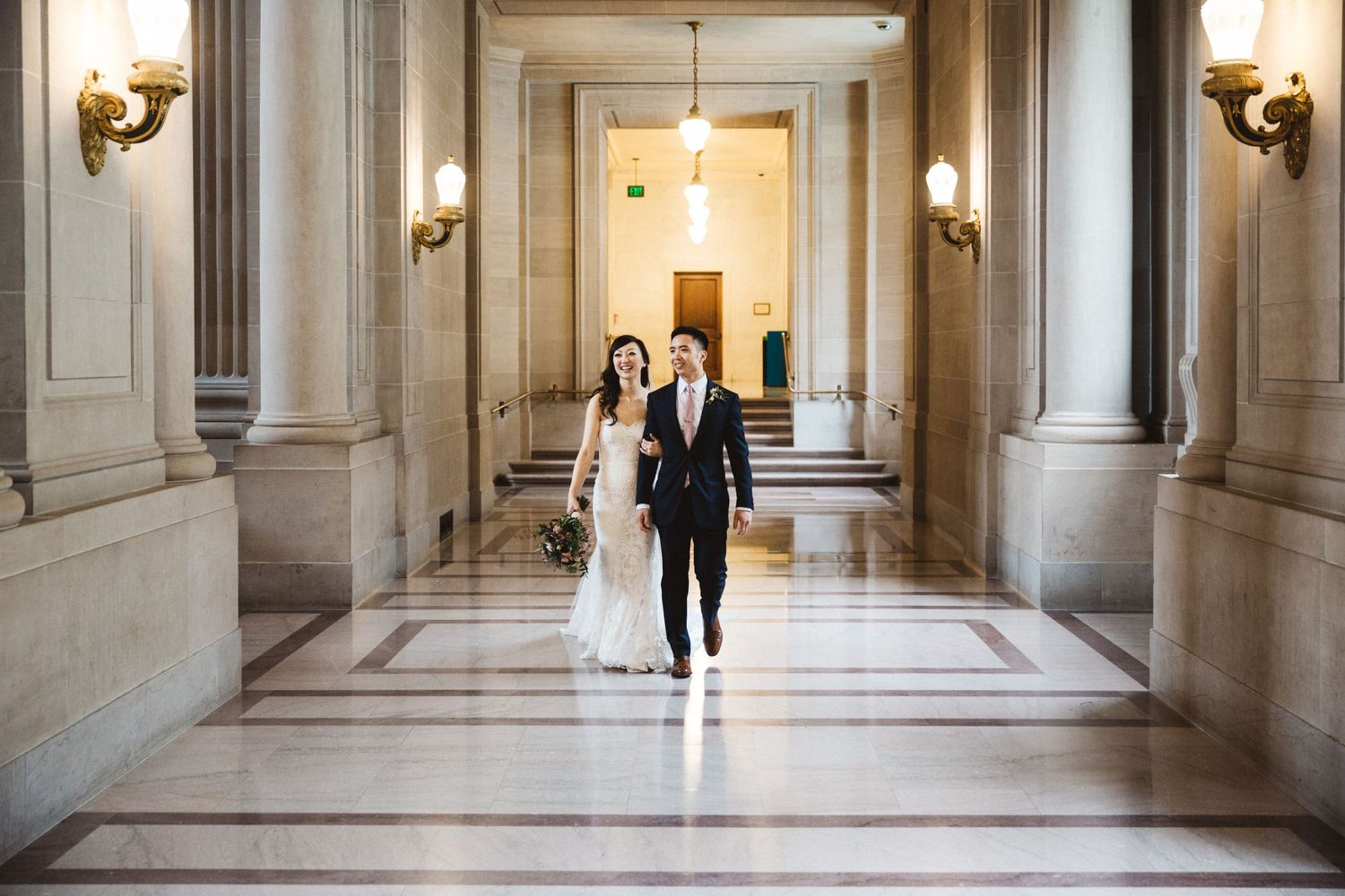 A couple walks through a bright marble corridor lined with classical columns and wall sconces.