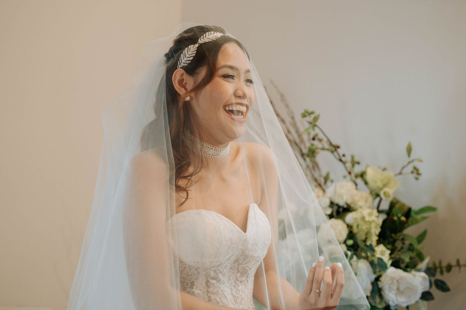 A bride wearing a lace wedding dress and veil smiles joyfully indoors.