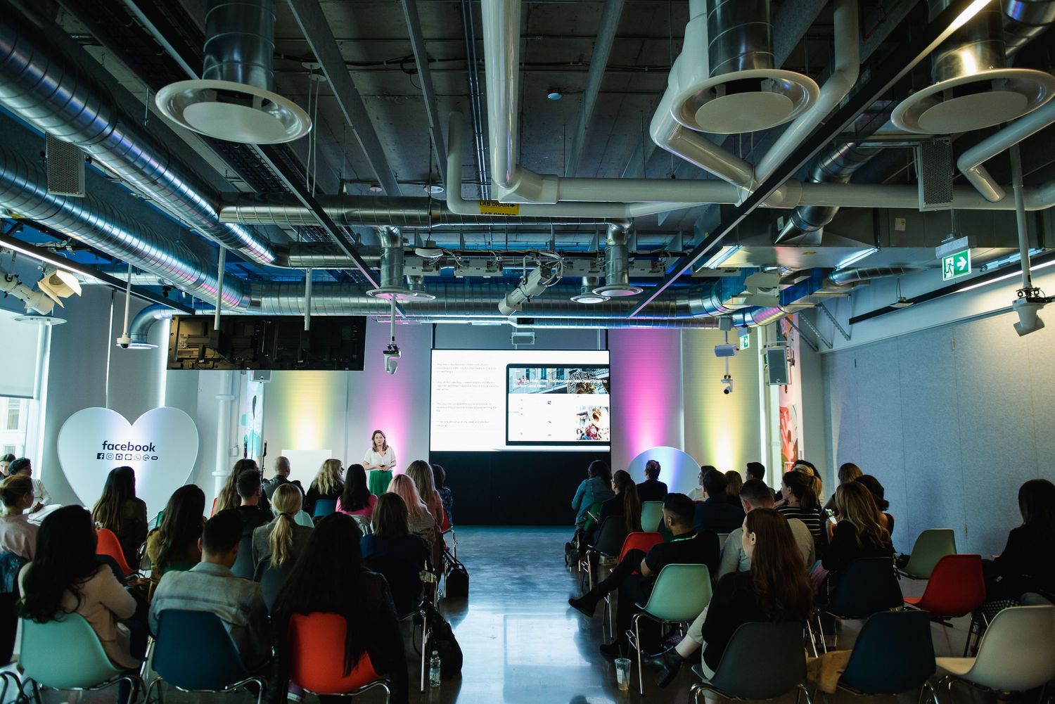Conference attendees seated in rows at modern event space with industrial ceiling and colored lighting.