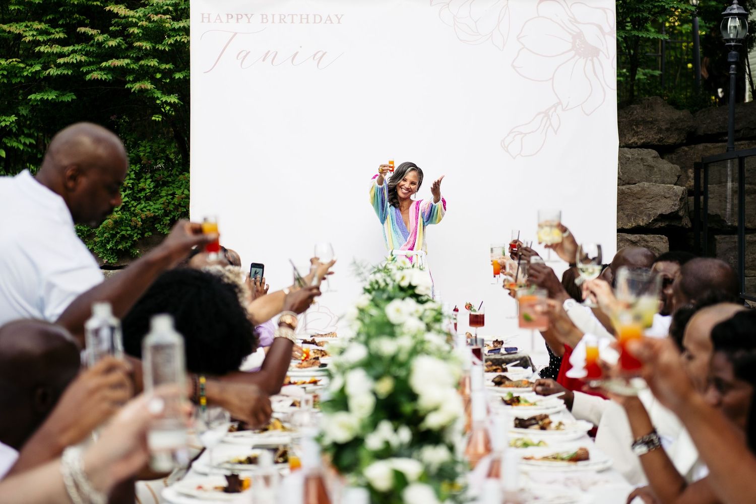 Guests raise their glasses in celebration at an outdoor wedding reception with floral centerpieces.