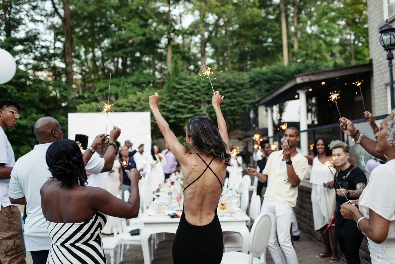 People dancing and celebrating at an outdoor party with white tables and string lights.