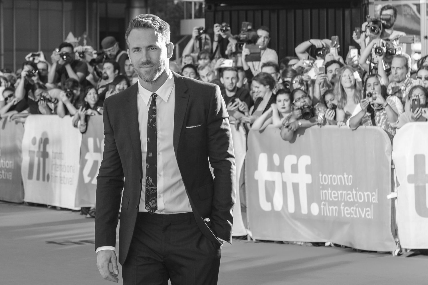 Man in black suit walks confidently on red carpet with TIFF backdrop and crowds.