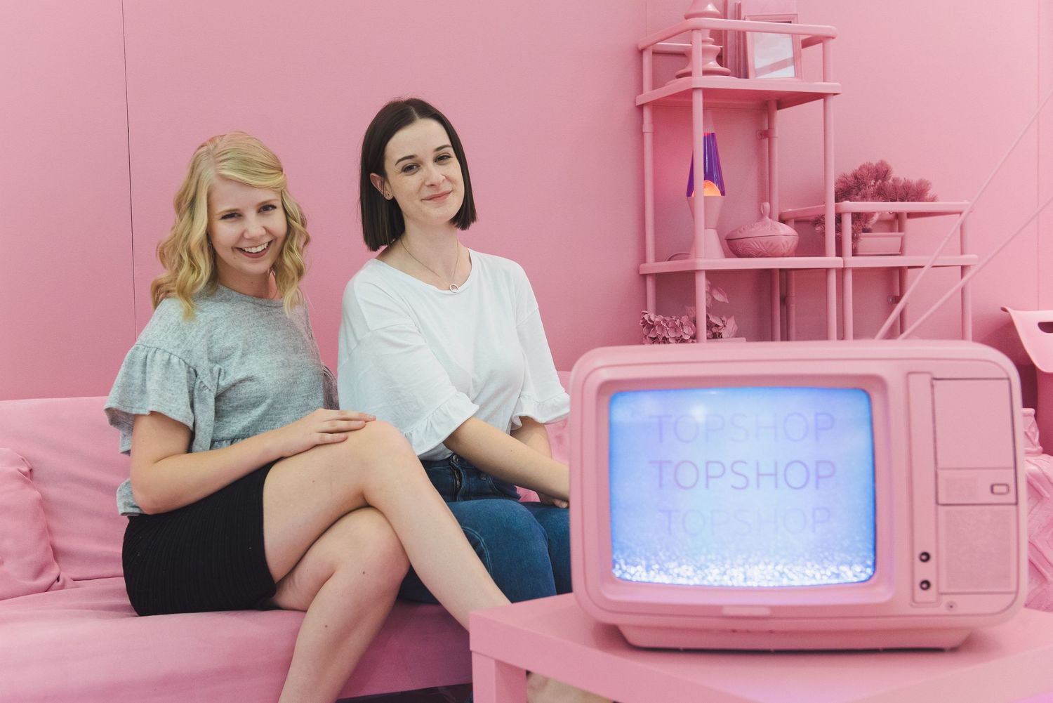 Two women sit on pink couch with vintage television in all-pink room setting.