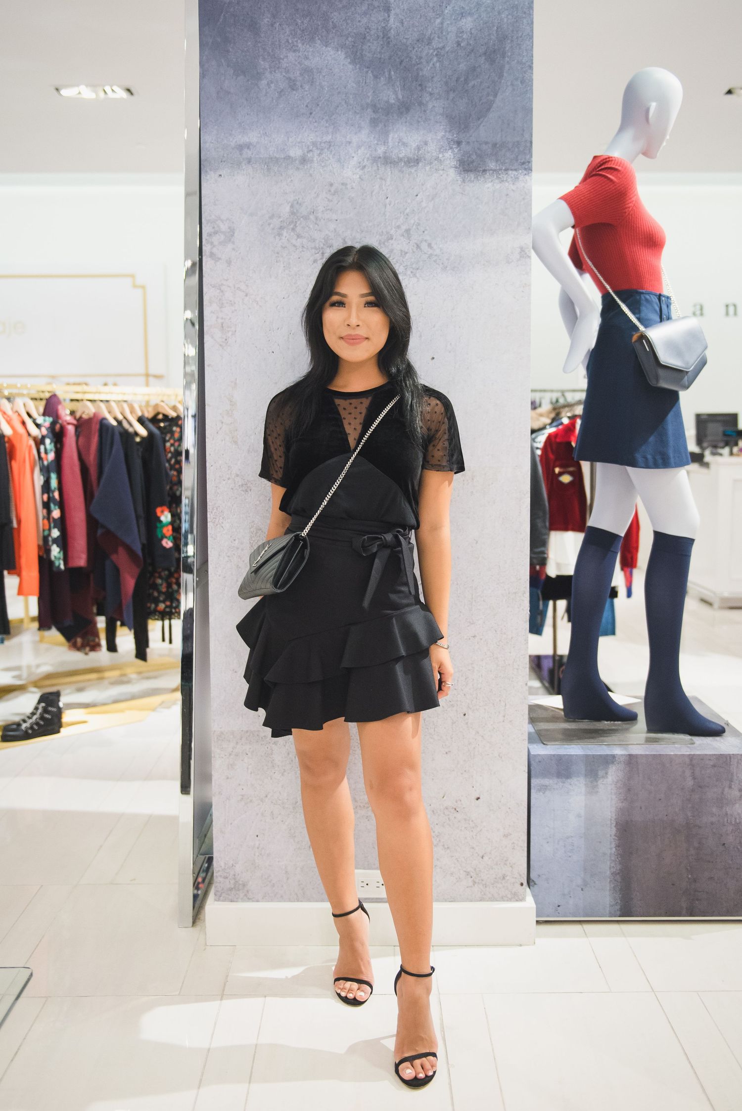 Stylish black ruffled dress with sheer sleeves shown in a clothing store with mannequins and racks in background.