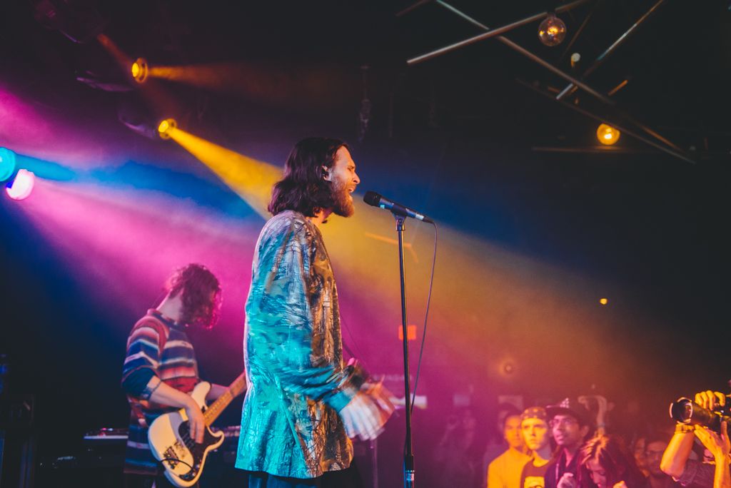 A performer sings on stage with colorful lighting and a crowd visible in the background.