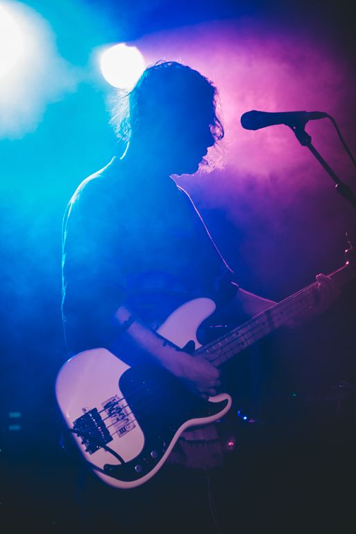 A silhouetted musician plays bass guitar against colorful blue and purple stage lighting.