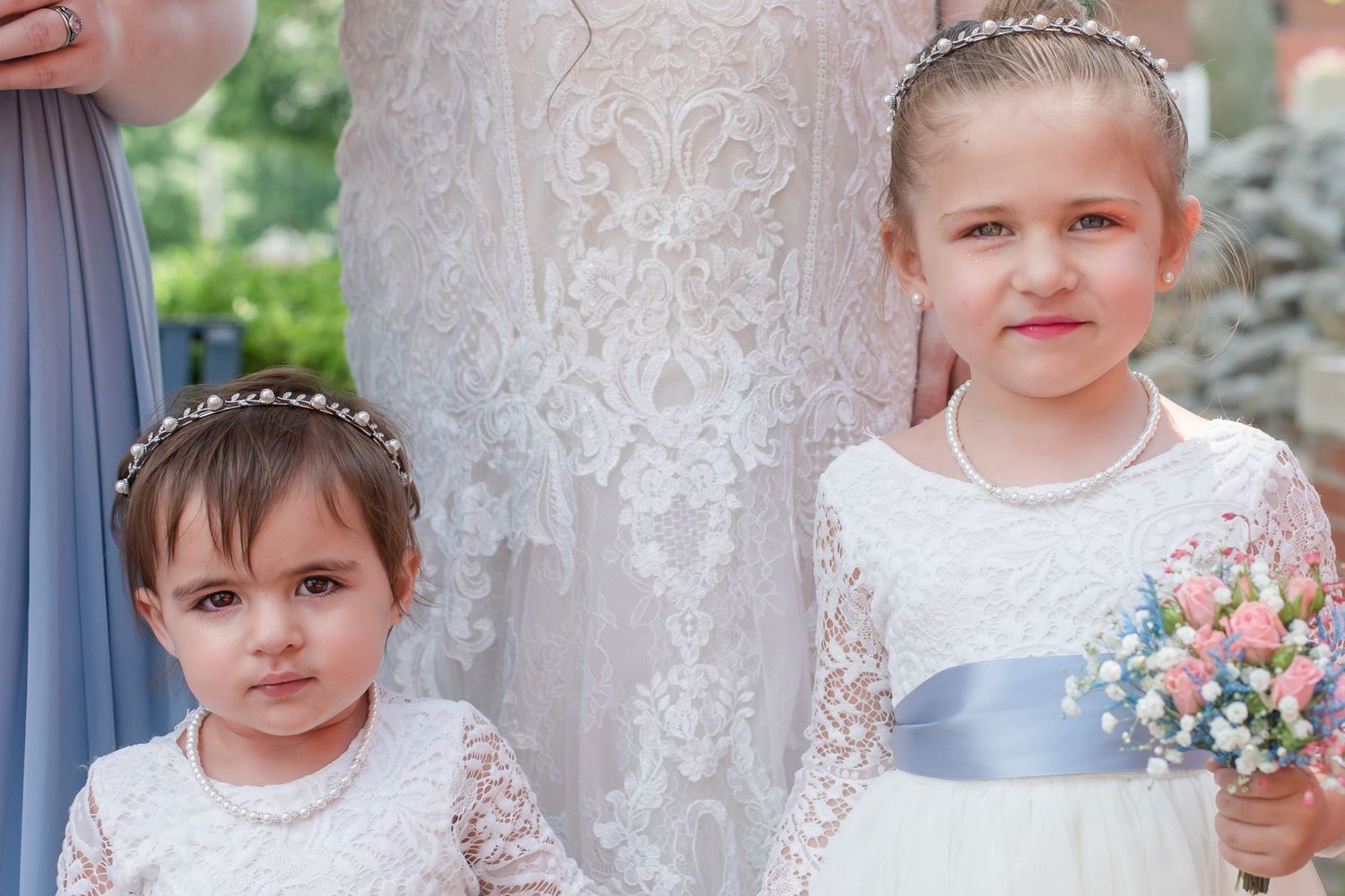 Two flower girls in white dresses with blue sashes stand beside a lace wedding gown.