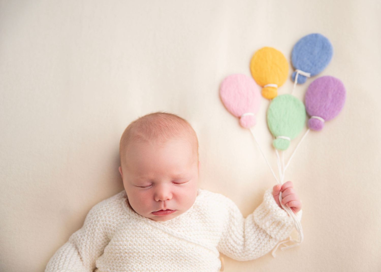 Sleeping newborn baby in white knit outfit posed with colorful felt balloon props.