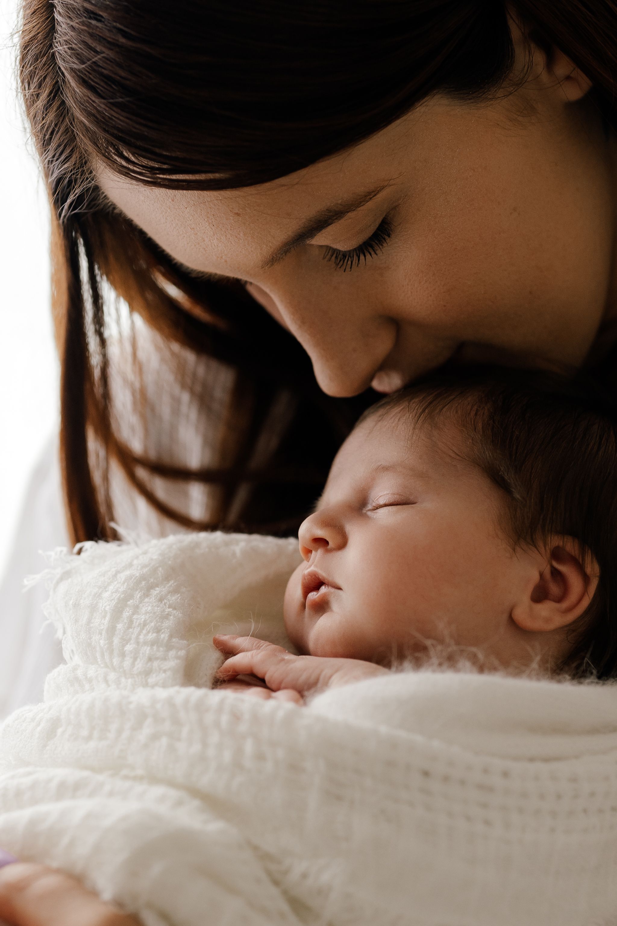 Loving mother with dark hair gently kissing her sleeping newborn baby wrapped in white blanket.