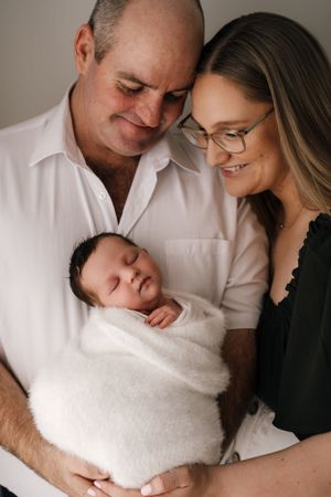 New parents lovingly gaze at their sleeping swaddled newborn baby in a tender studio portrait.