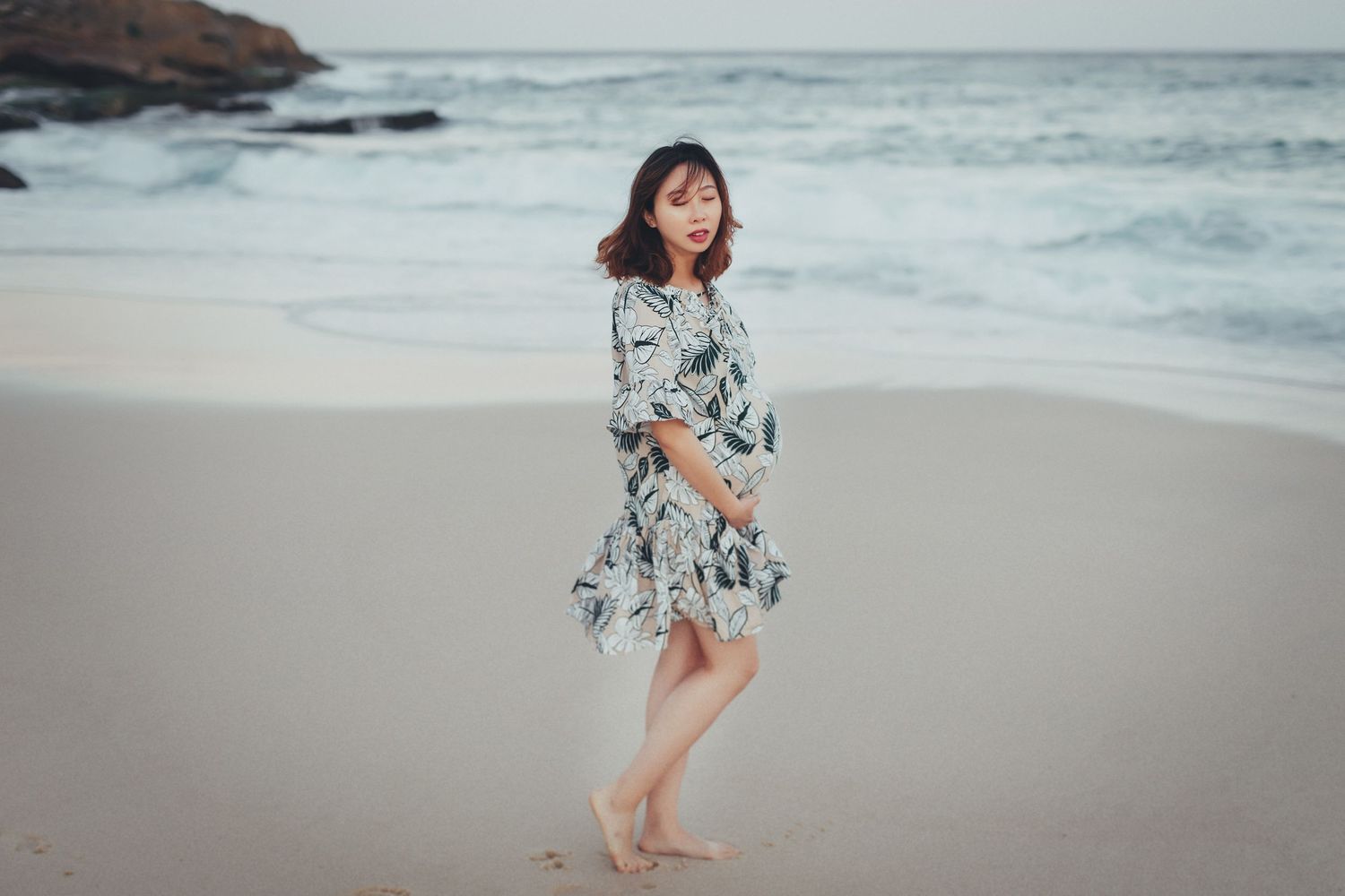 Pregnant woman in floral dress walking barefoot on sandy beach with ocean waves in background.