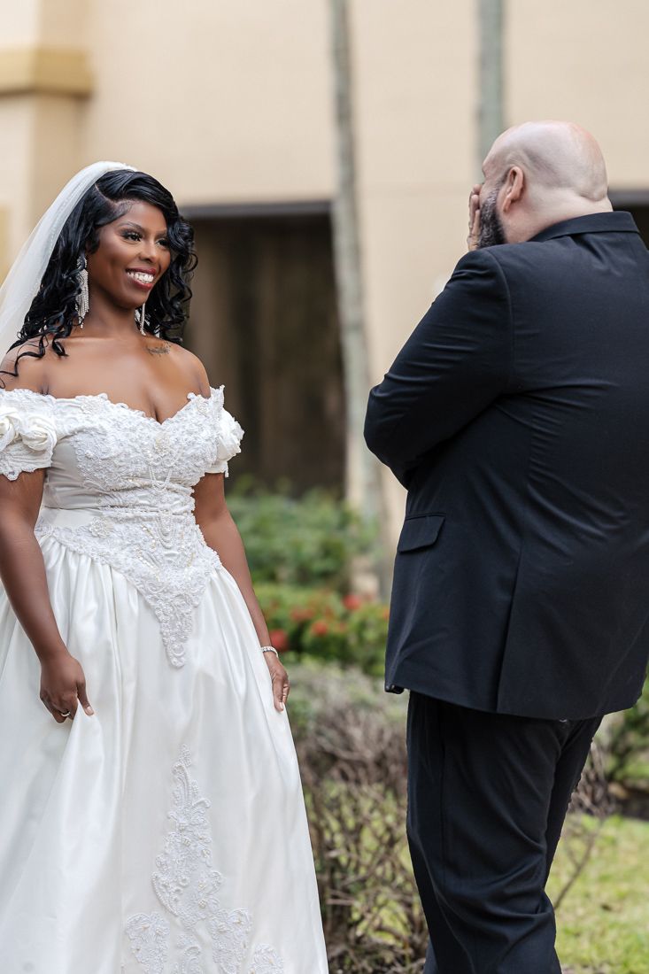 Bride in off-shoulder lace gown smiles during first look with groom at outdoor wedding venue.