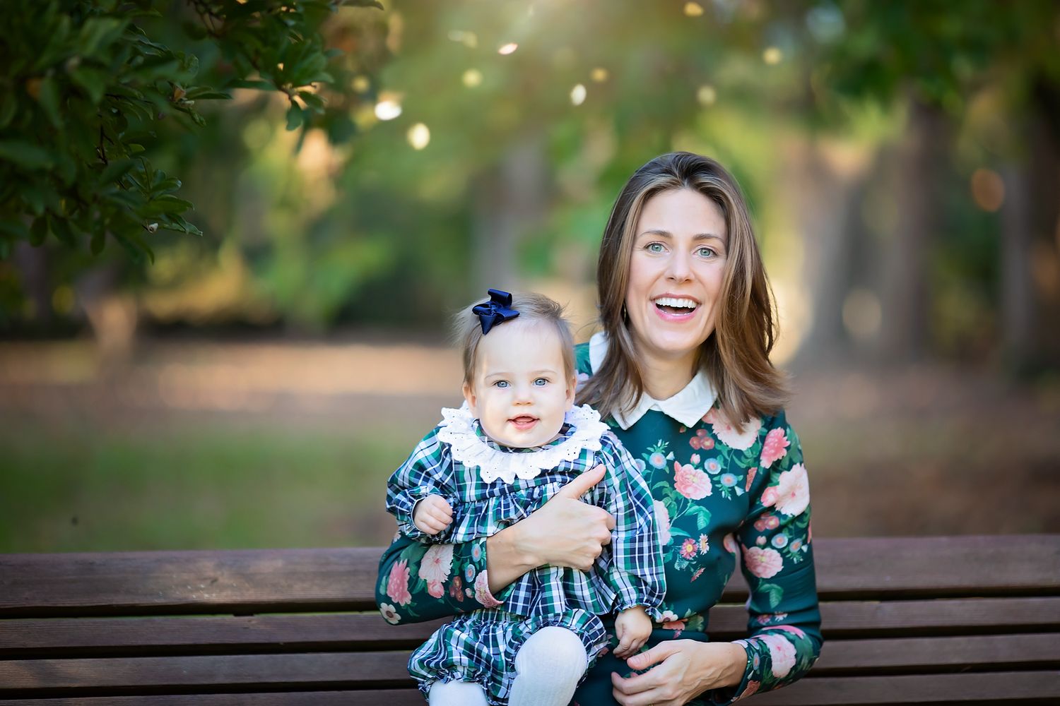 Smiling mother holding baby girl on a wooden bench in a sunny outdoor park setting.