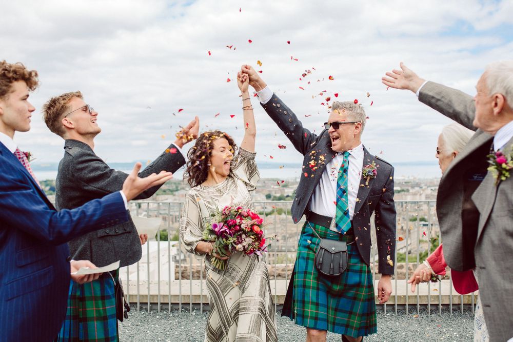 smiling bride and groom put arms in the air as confetti is thrown on them by their family on Calton Hill, Edinburgh