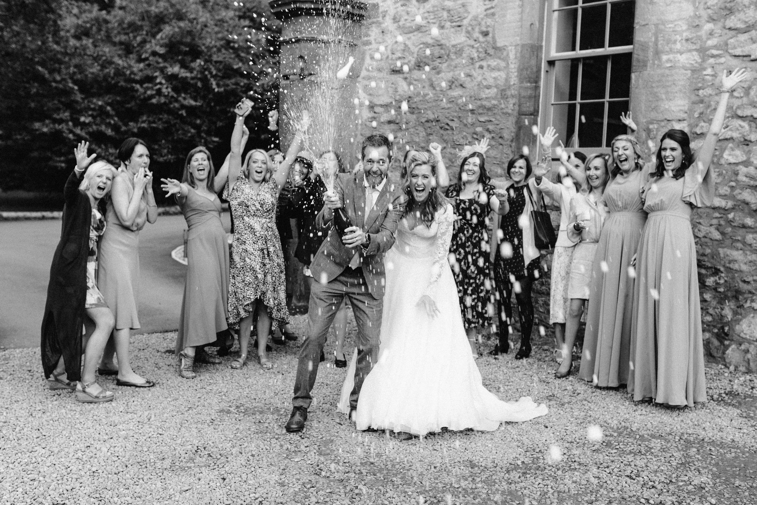 Wedding party and guests gather outside a rustic stone building for group photos.