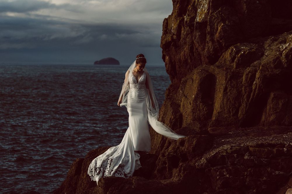 Bride stood on cliffs by the North sea, in evening sun and Bass Rock behind