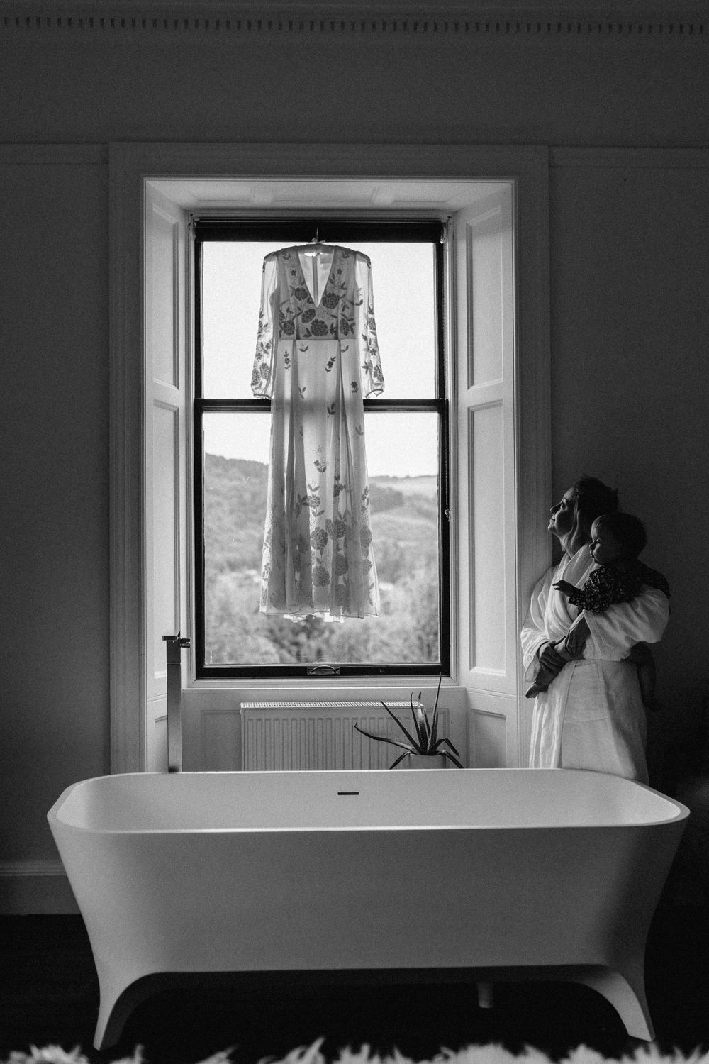Beautiful wedding dress hanging in the window with bride and her baby in arms looking up at dress.