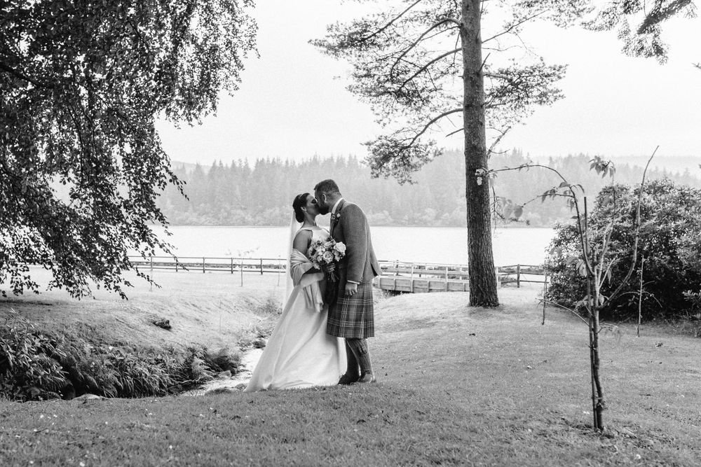 Bride and groom kiss under tress by a lake