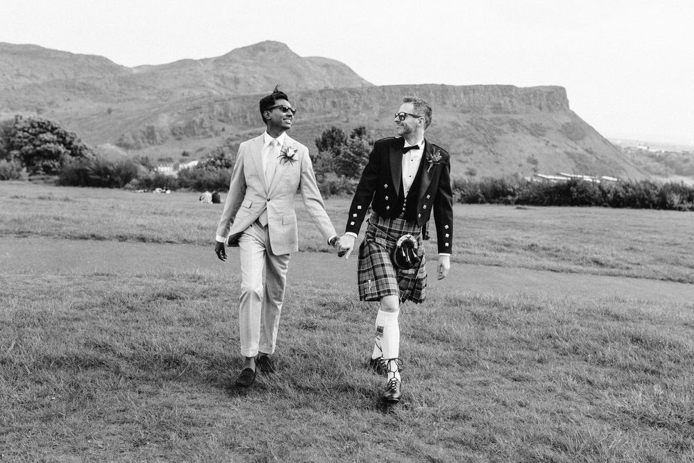 Two grooms walking and laughing together whilst holding hands in front of Arthurs seat in Edinburgh, Scotland