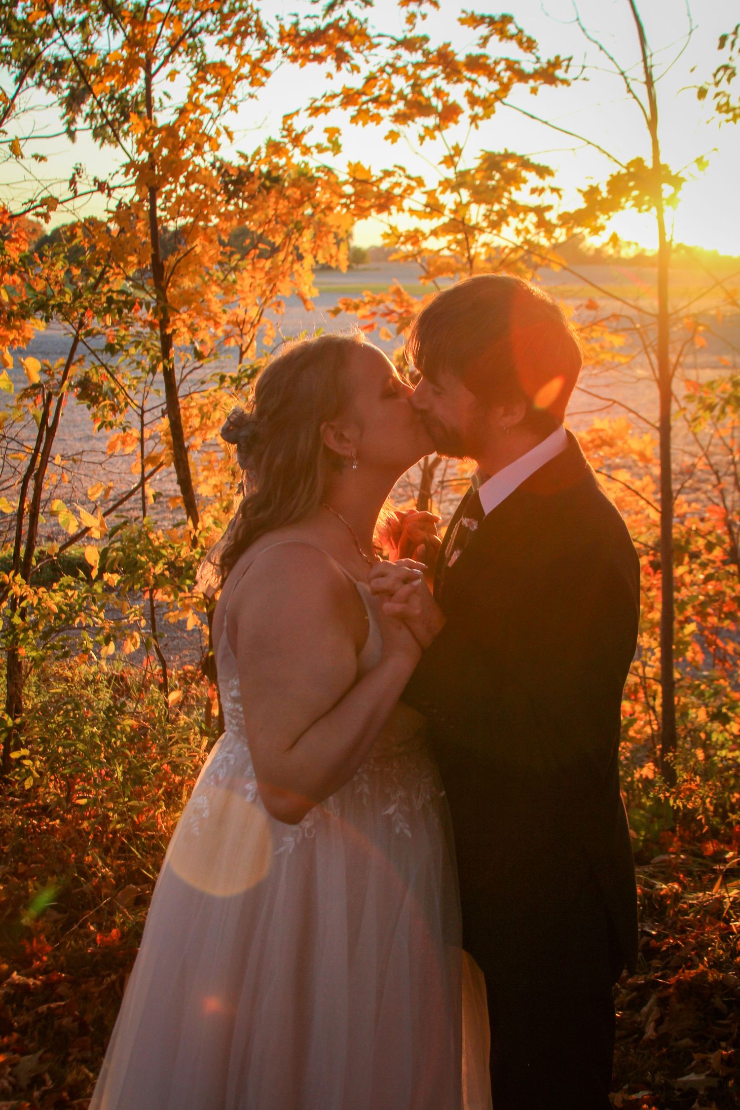 A romantic wedding couple shares a kiss at sunset with autumn foliage and golden light in the background.
