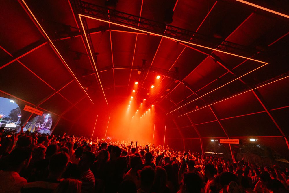 Packed concert venue illuminated by red lighting and laser effects during a performance.